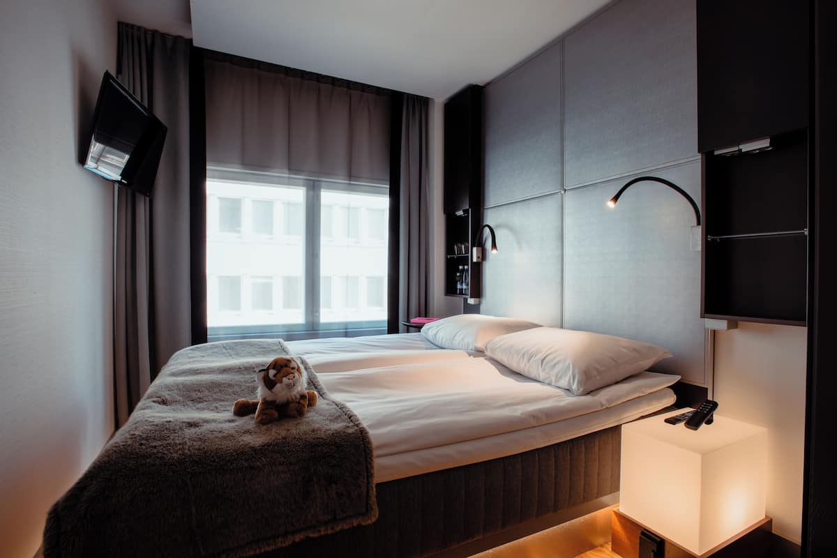 Una habitación de hotel moderna con una cama grande, un televisor montado en la pared, una lámpara de noche y un ventanal con cortinas.