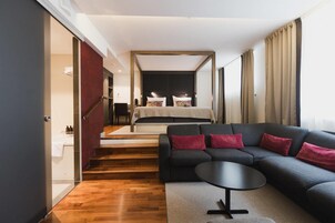 Una habitación de hotel moderna con una cama grande, un sofá oscuro, una mesita pequeña y una escalera que lleva a un baño.