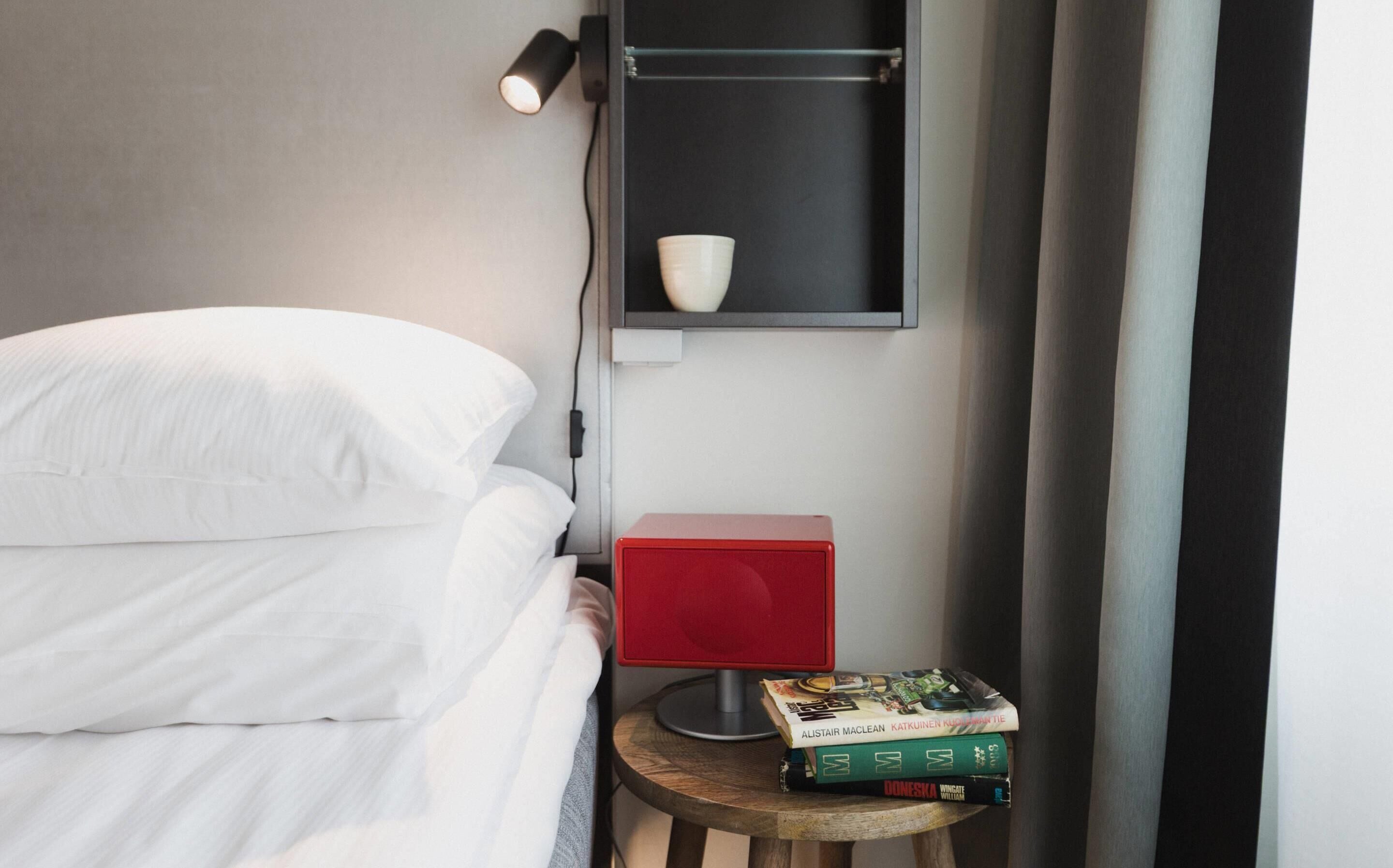 A neatly made bed with white linens, a red speaker, and a small table with books.