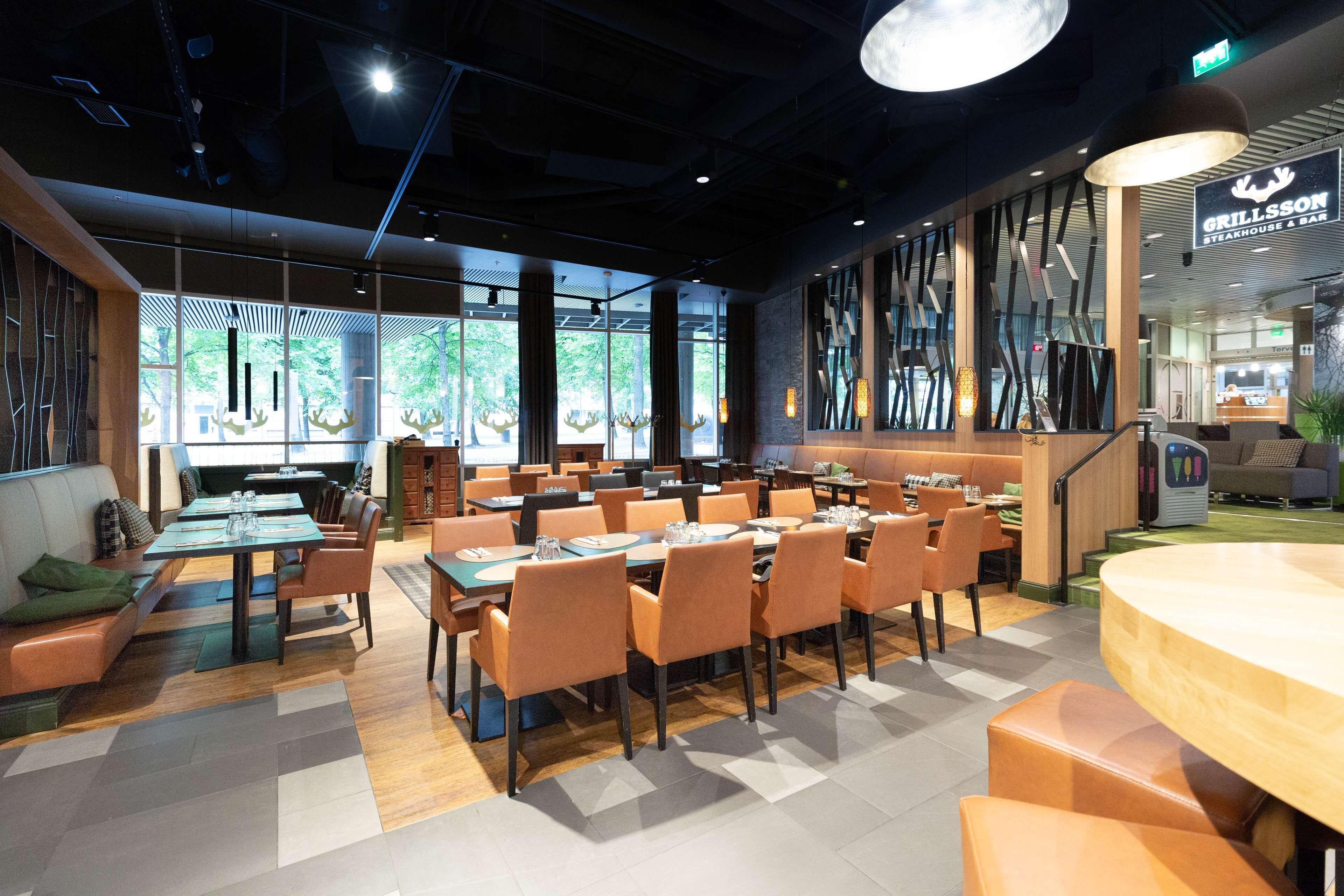 Un restaurant moderne doté de nombreuses tables et chaises, de grandes fenêtres et d’un espace grill visible.