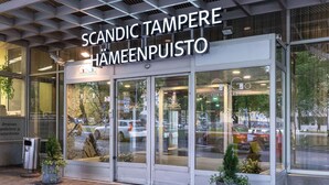 L’entrée d’un hôtel avec une façade en verre, une enseigne indiquant « Scandinavic Tampere Hämmenpisto » et des plantes en pot à l’extérieur.
