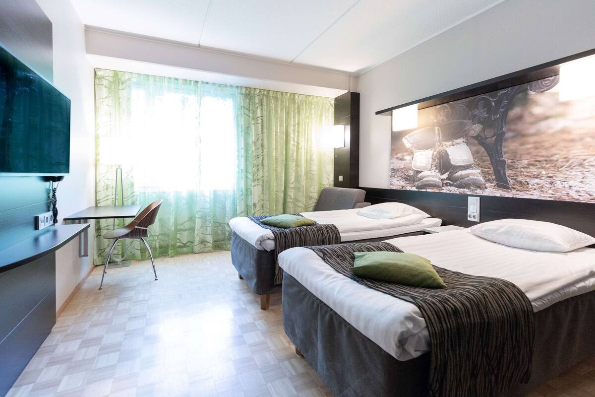 Ein Hotelzimmer mit zwei Betten, einem Schreibtisch, einem Stuhl und einem großen Fenster mit Vorhängen.