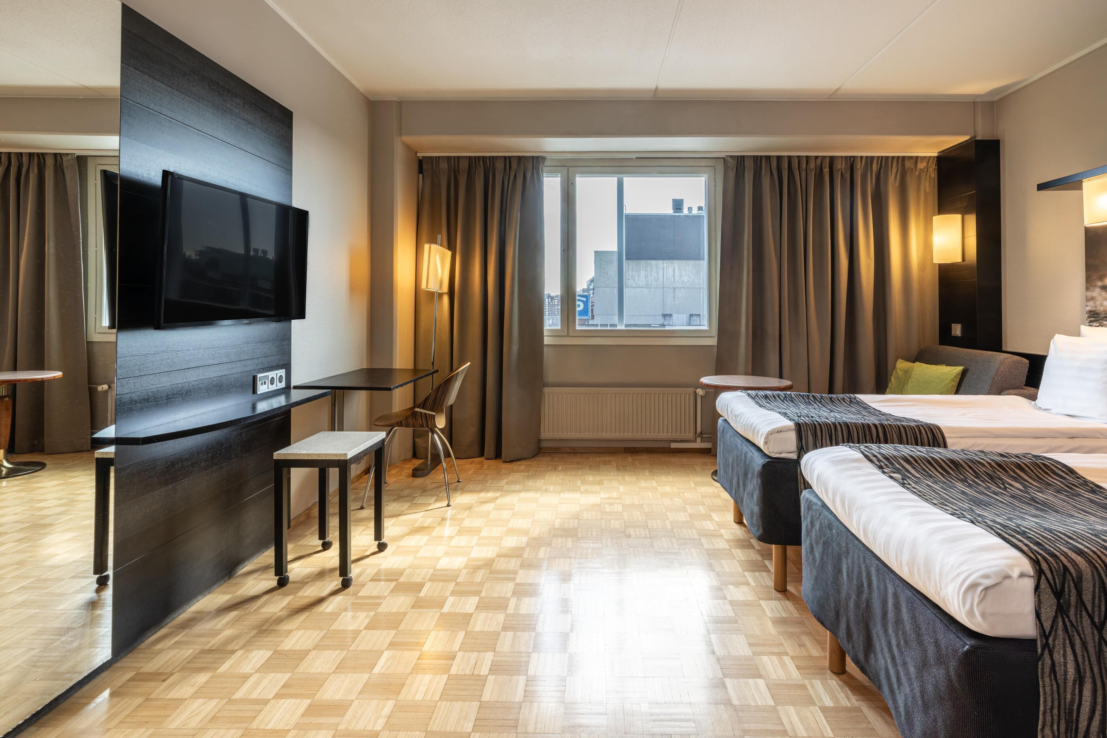 Et hotelværelse med et fladskærms-tv, et skrivebord, to senge, et vindue med gardiner og udsigt til bygninger.