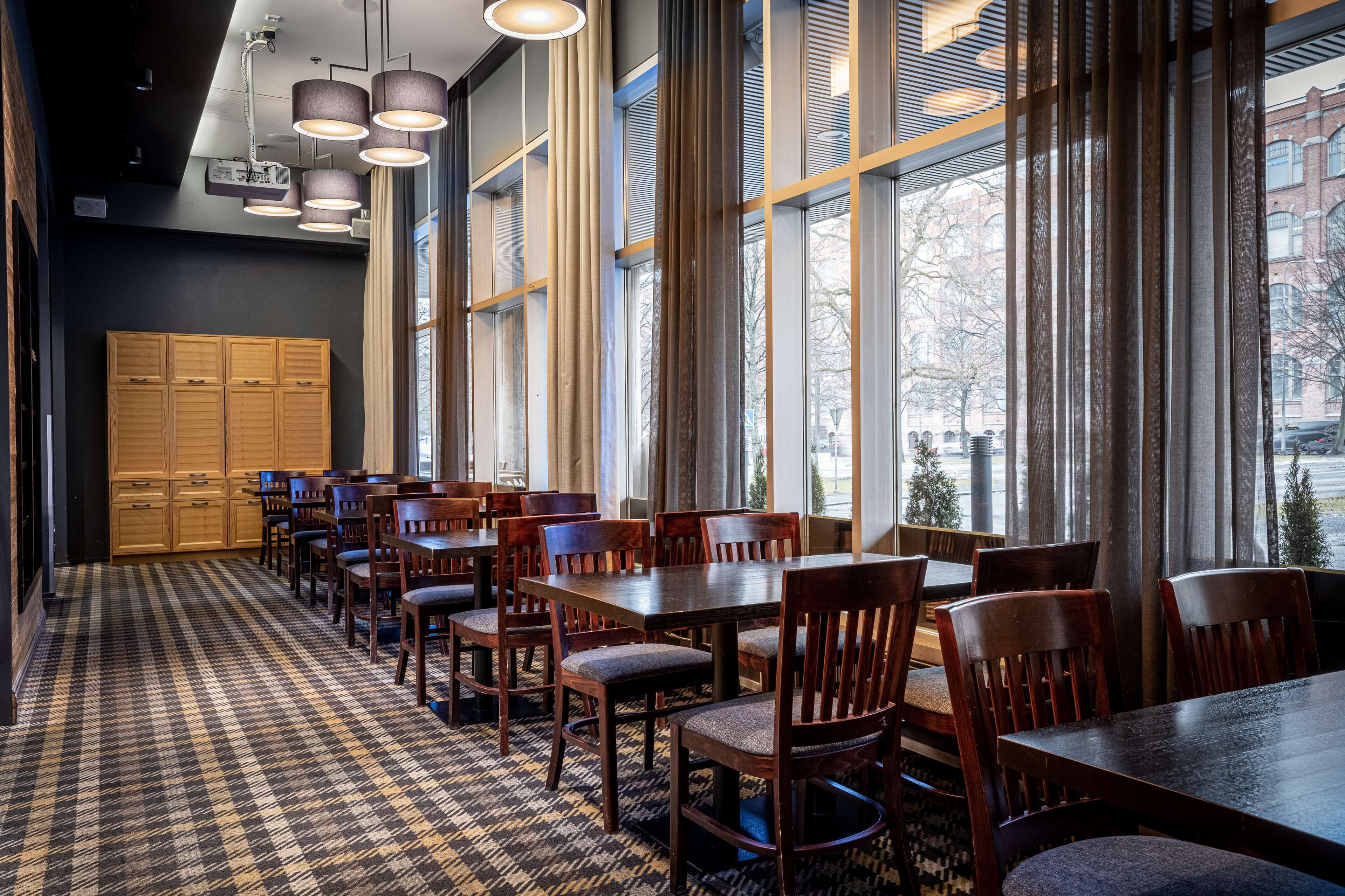A dining area with wooden tables and chairs, large windows with blinds, and a patterned carpet.