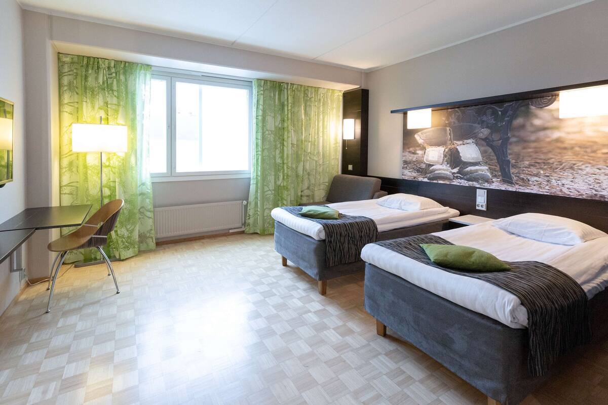 Ein Hotelzimmer mit zwei Betten, einem Schreibtisch, einem Stuhl und einem großen Fenster mit Vorhängen.