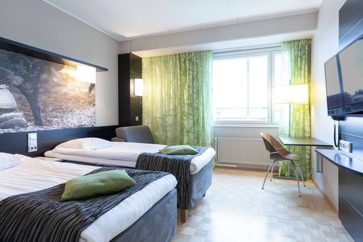 Ein Hotelzimmer mit zwei Betten, einem Schreibtisch, einem Sessel, einem Fernseher und einem Fenster mit Vorhängen.