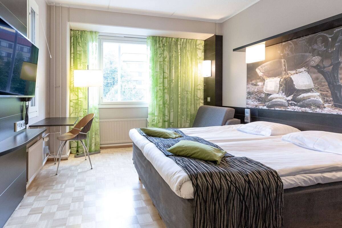 Ein Hotelzimmer mit einem Bett, einem Schreibtisch, einem Stuhl, einem fensterbelaubten Fenster und einem Bild an der Wand.