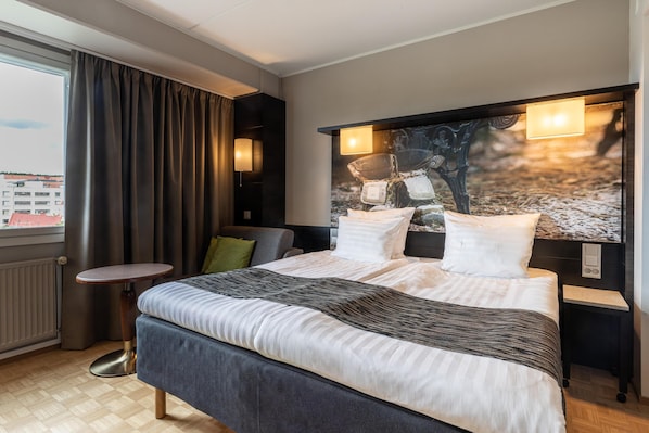 A hotel room with a large bed, a small round table, a view of the city, and a painting on the wall.