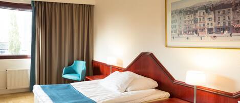 A hotel room with a bed, a chair, a window with curtains, a radiator, and a framed picture on the wall.