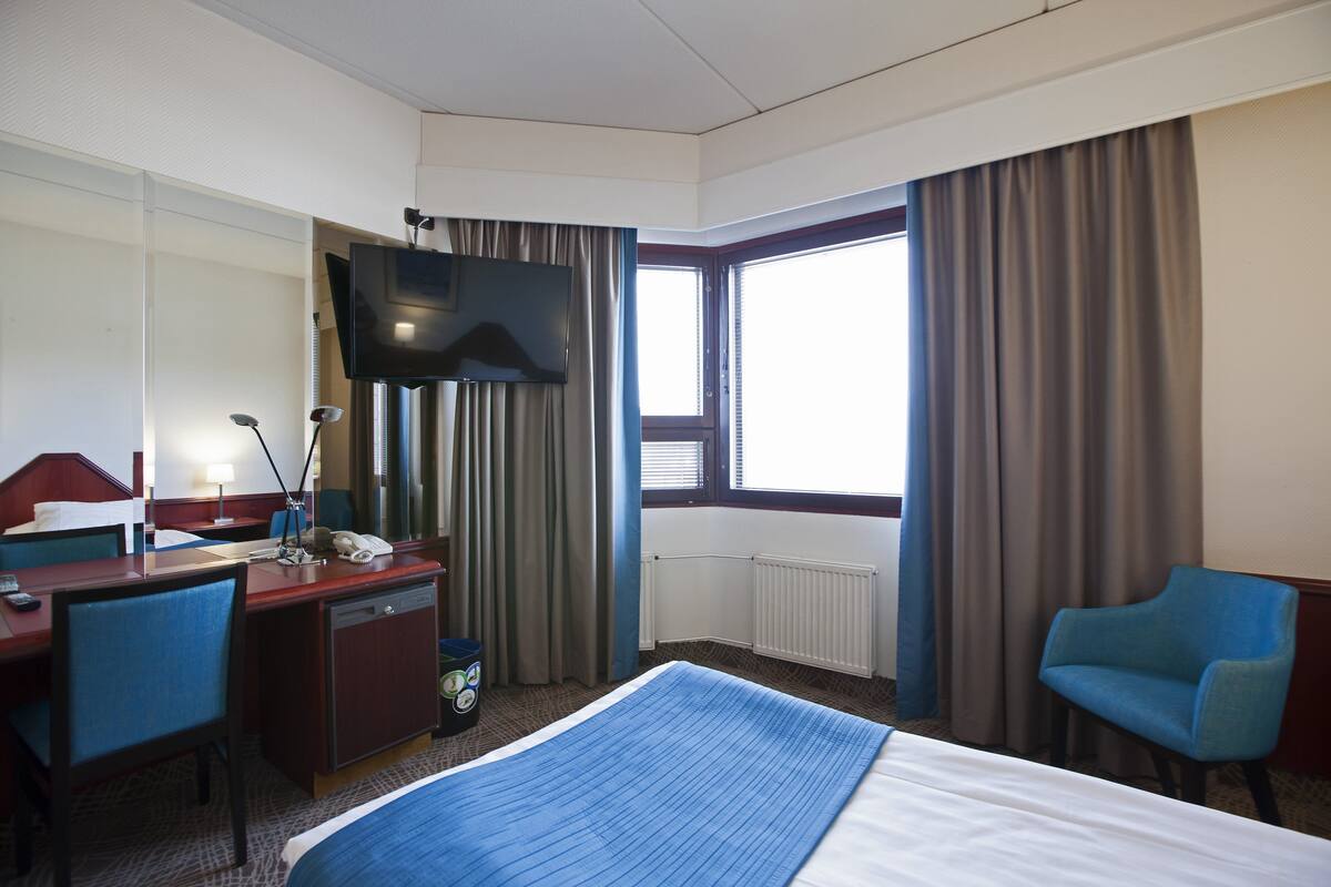A hotel room with a bed, a desk with a telephone, a chair, a television, and a window with curtains.