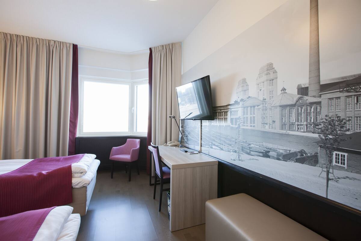 A hotel room with a bed, a desk, a chair, a TV, and a large cityscape painting on the wall.