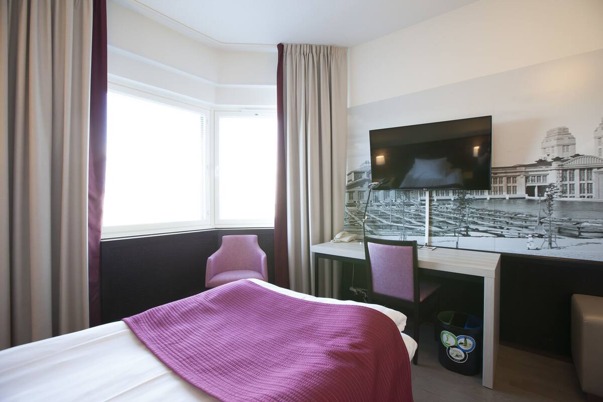 A hotel room with a bed, a desk with a computer, a television, and a window with curtains.