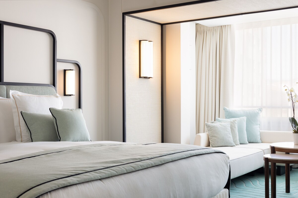 A modern hotel room with a large bed, a sofa, a small table, and a window with curtains.