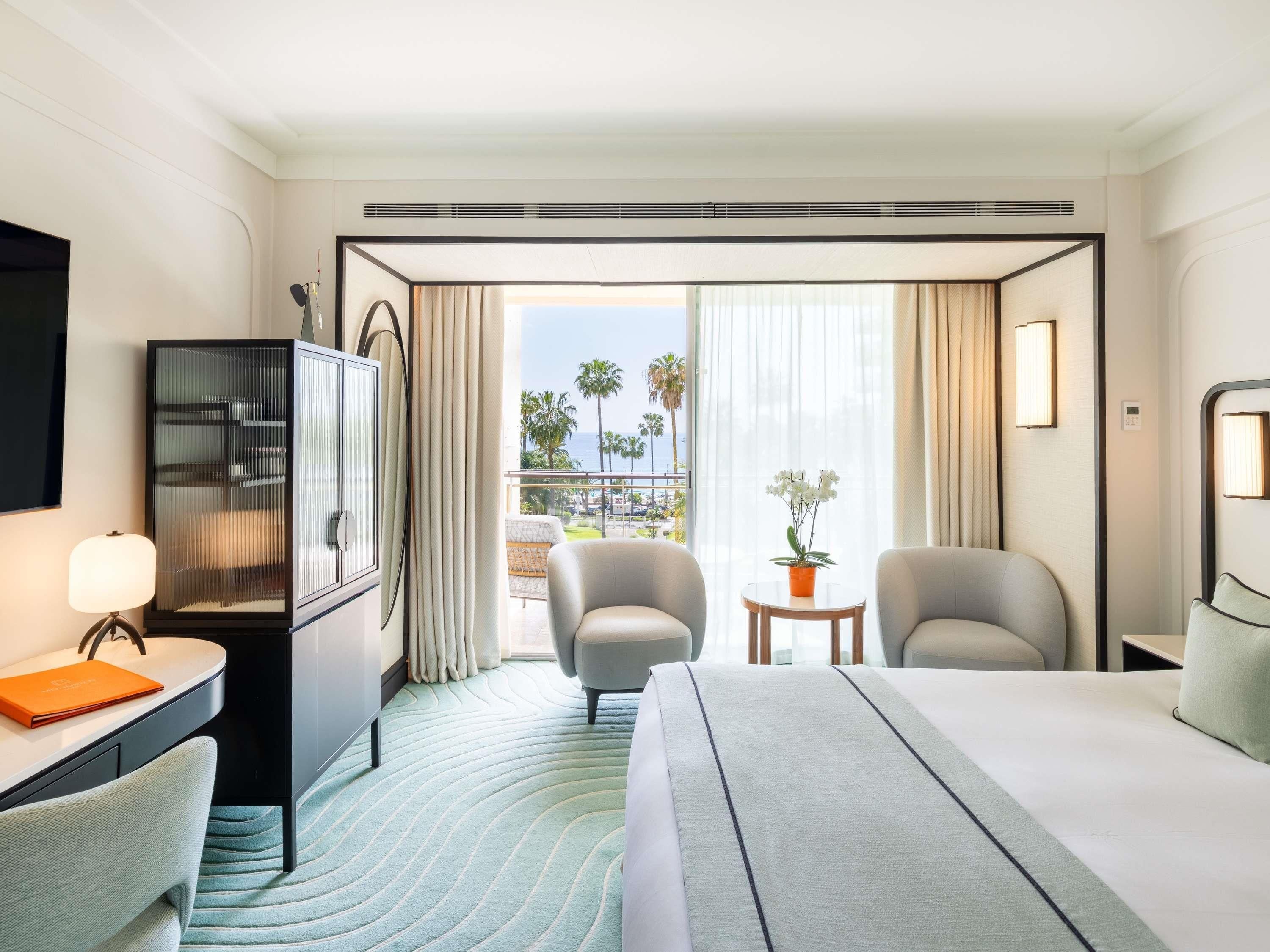Ein Hotelzimmer mit Bett, zwei Sesseln, einem Schreibtisch mit Lampe, einem Fernseher, einer Minibar und Blick auf Palmen und einen Pool.
