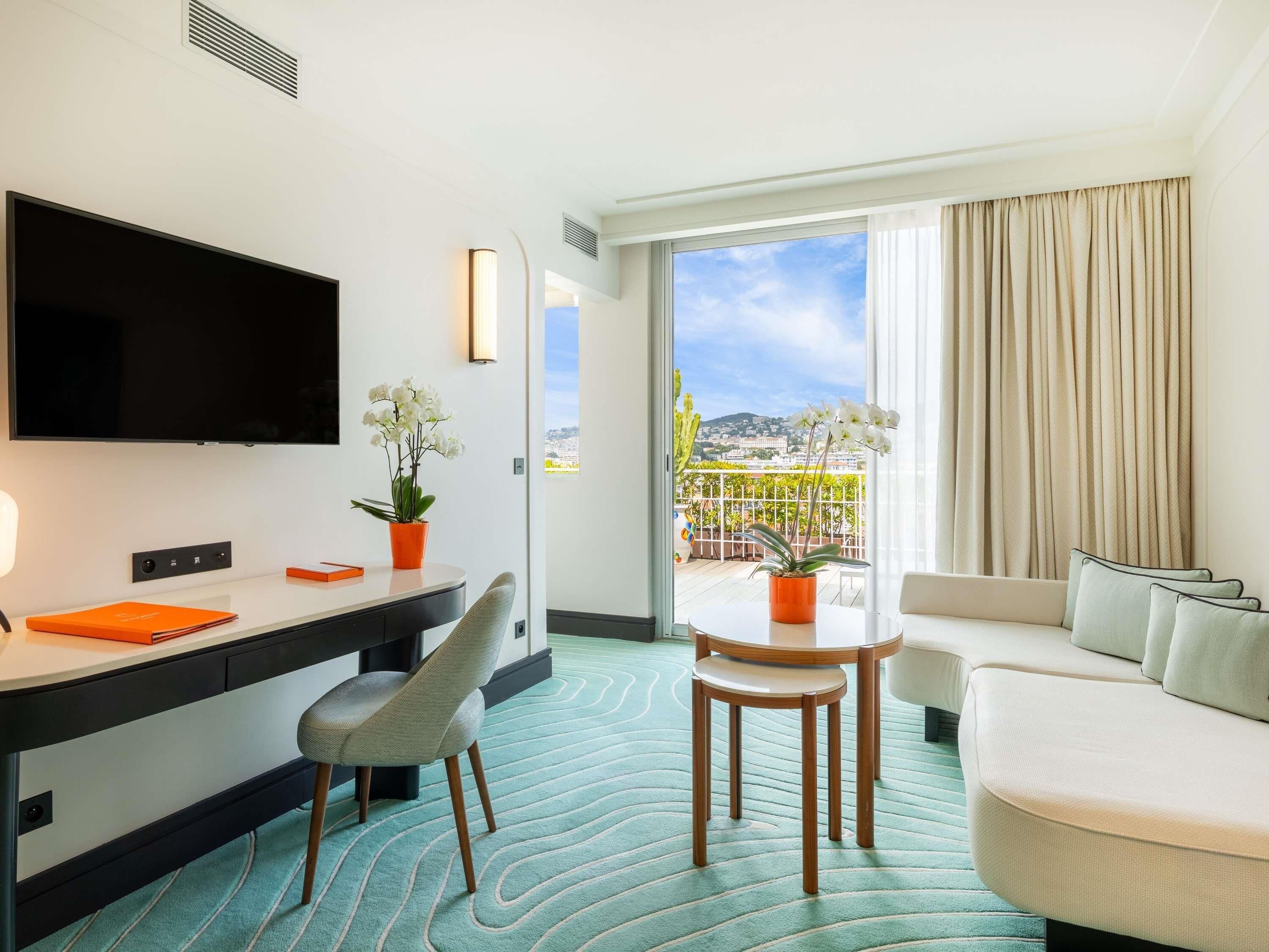 Habitación de hotel con televisión de pantalla plana, un escritorio con silla, una mesa de comedor con sillas, un sofá y un balcón con vista a la ciudad.