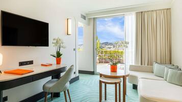 Habitación de hotel con televisión de pantalla plana, un escritorio con silla, una mesa de comedor con sillas, un sofá y un balcón con vista a la ciudad.