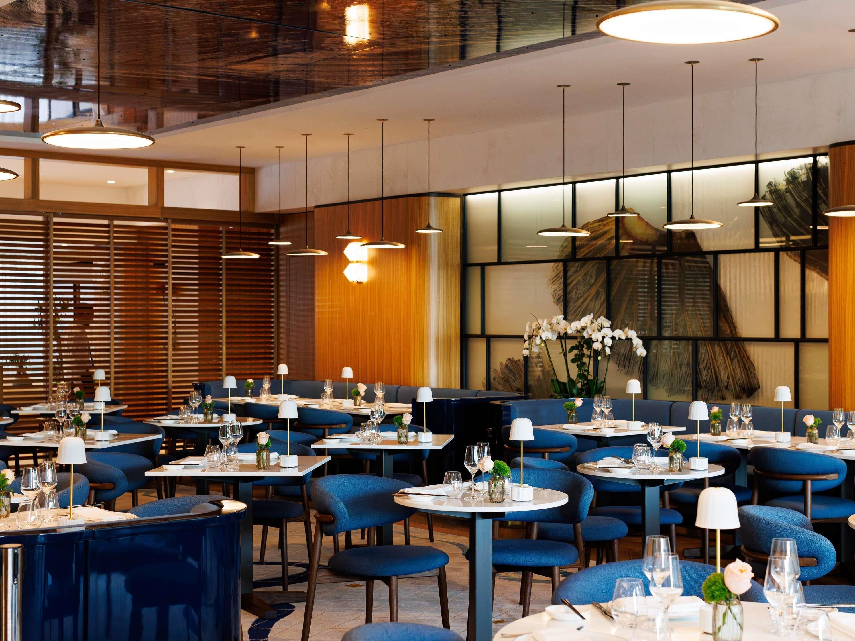 A modern restaurant interior with round tables, blue upholstered chairs, and pendant lighting.