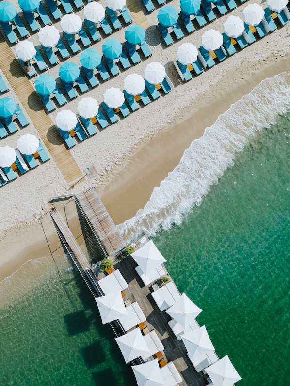 Vista aérea de una playa con filas de tumbonas y sombrillas, un muelle de madera que conduce al agua y un mar turquesa y transparente.