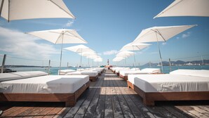 A row of wooden beds with white linens and white umbrellas, leading to a beach with boats and a clear sky. - Mondrian Cannes (Cannes)