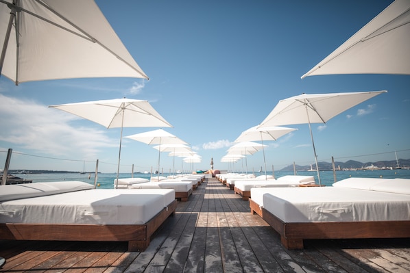 A row of wooden beds with white linens and white umbrellas, leading to a beach with boats and a clear sky. - Mondrian Cannes (Cannes)