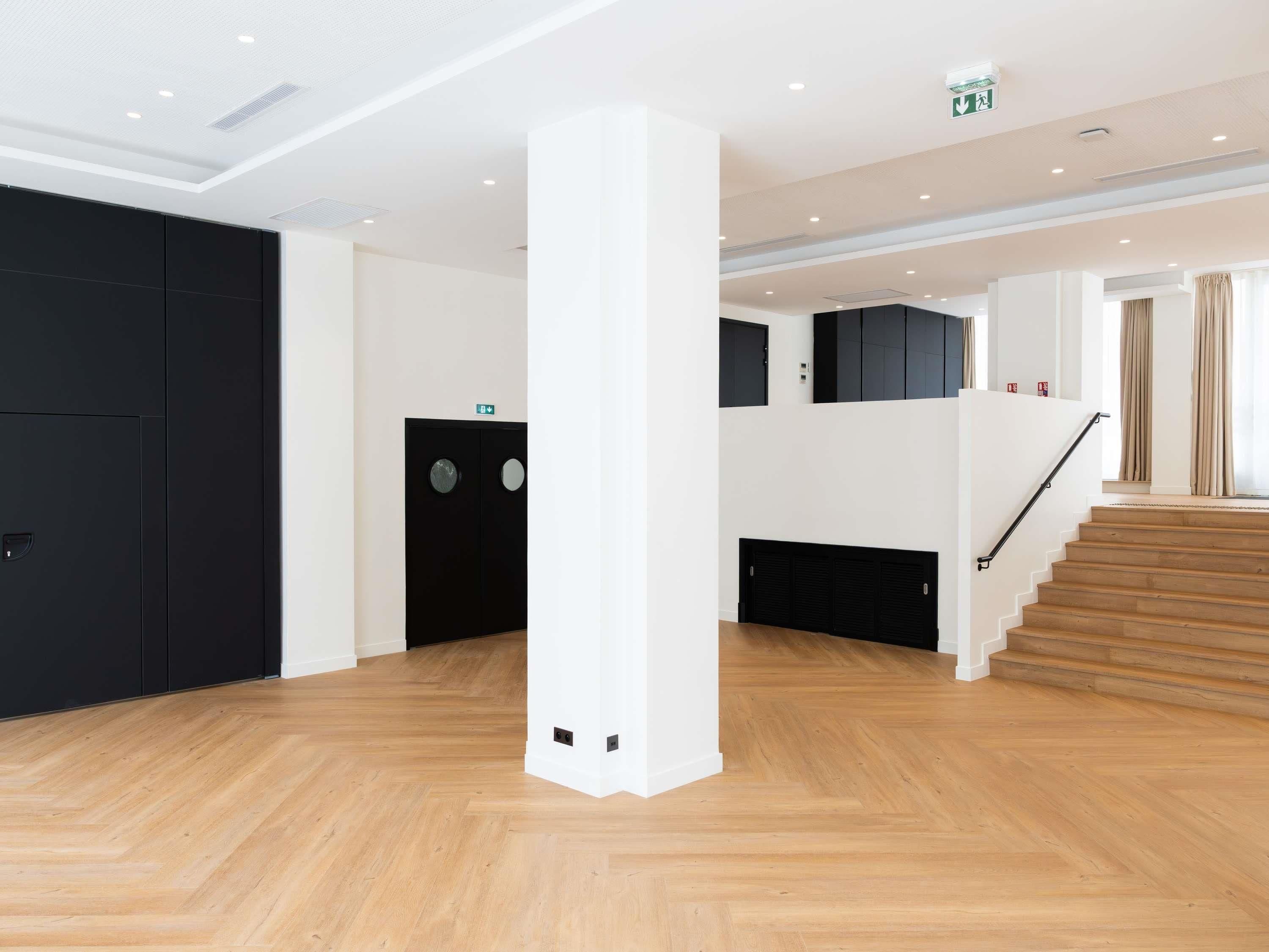A spacious hotel lobby with a wooden floor, a staircase, and a black elevator.