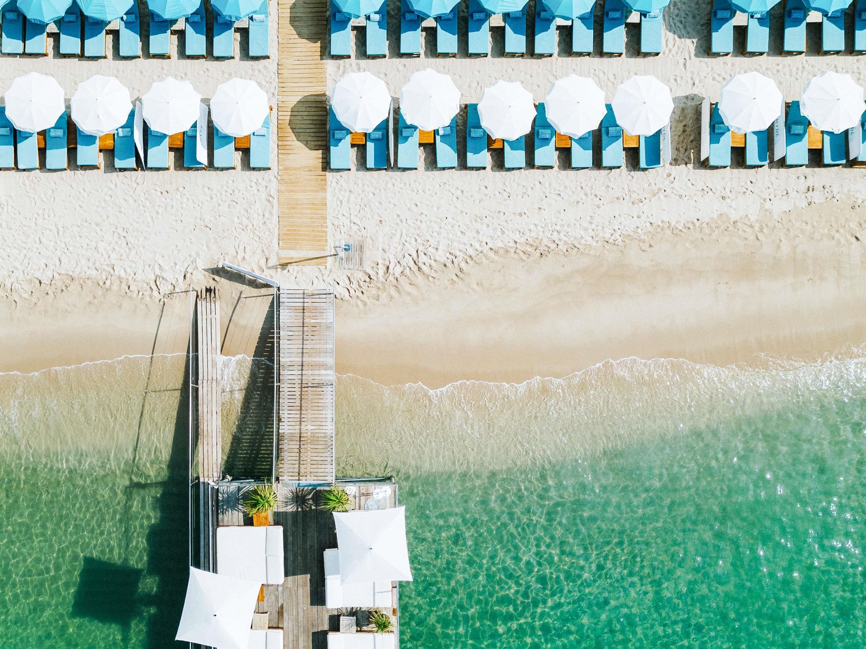 Vista aérea de un resort frente a la playa, con un paseo de madera que conduce a una zona de piscina, rodeada de sombrillas y sillas de playa.