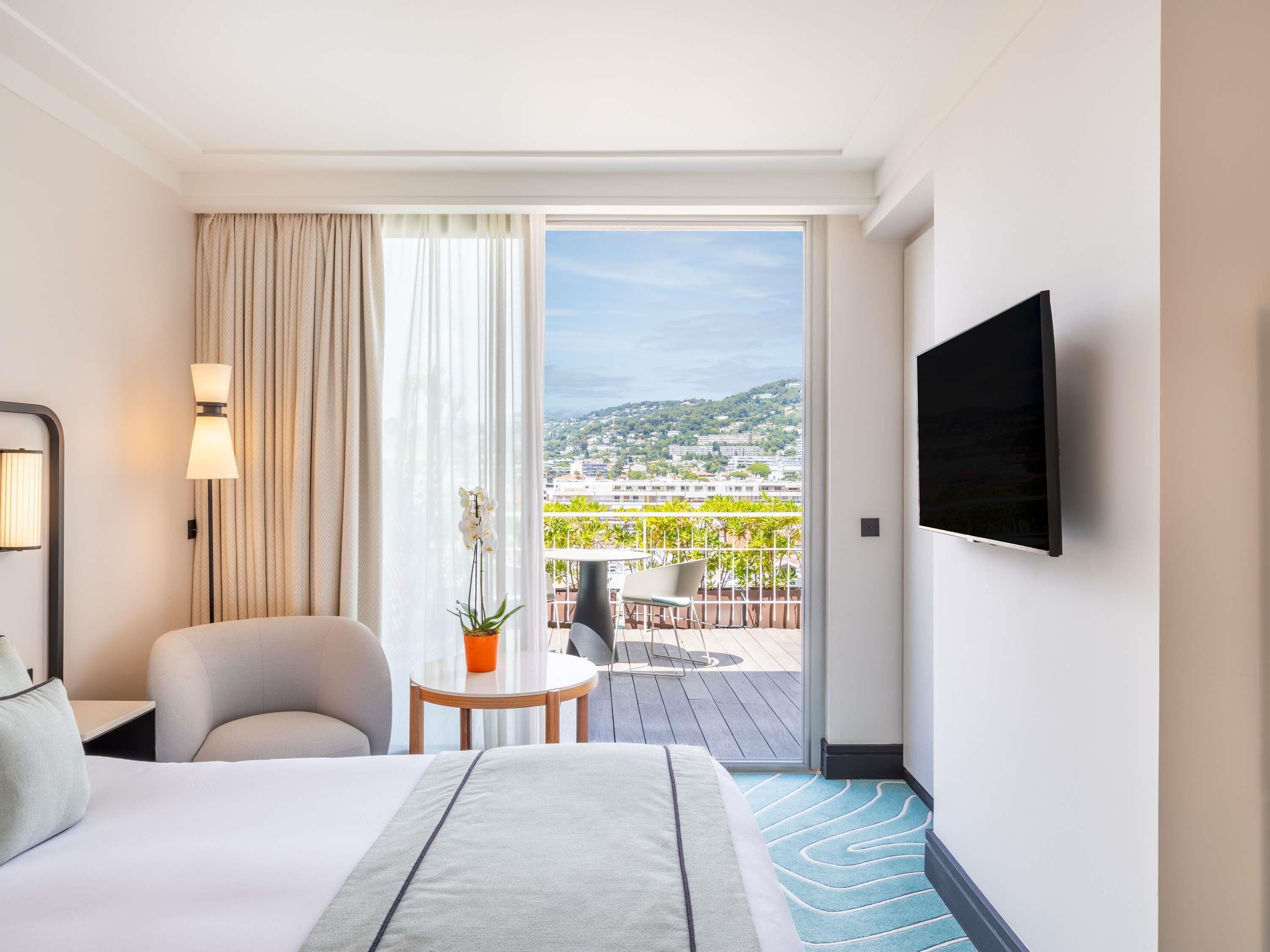 Een hotelkamer met een bed, een stoel, een kleintje tafeltje met een plant, een televisie en een balkon met uitzicht op de stad.