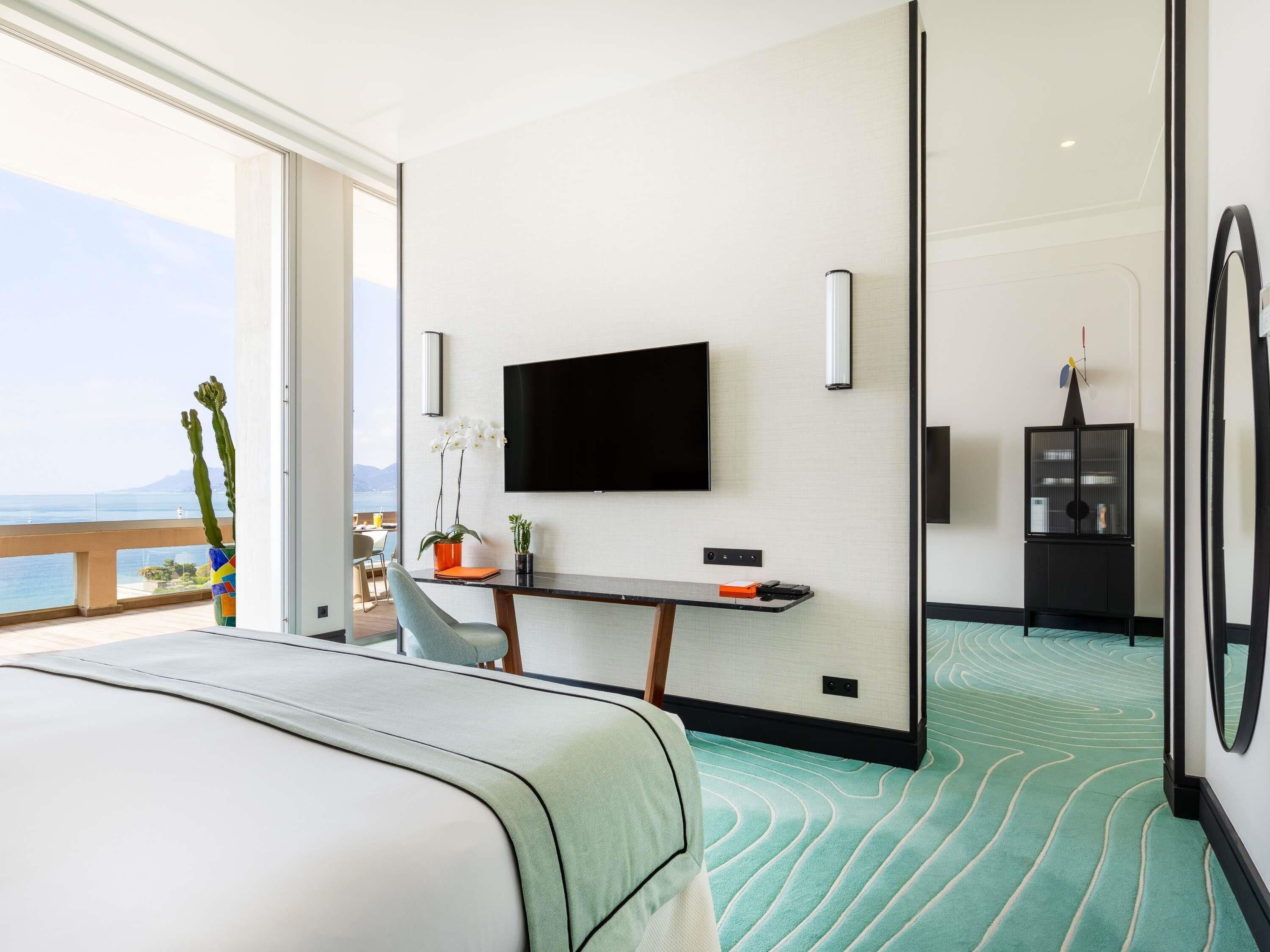 Een hotelkamer met een bed, een bureau met een televisie, een plant en uitzicht op zee.