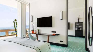 Een hotelkamer met een bed, een bureau met een televisie, een plant en uitzicht op zee.