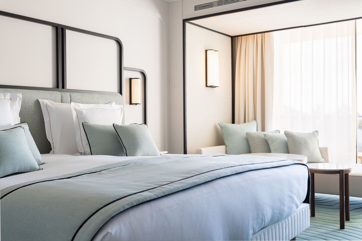 A modern hotel room with a large bed, a small table, and a view of the city through large windows.