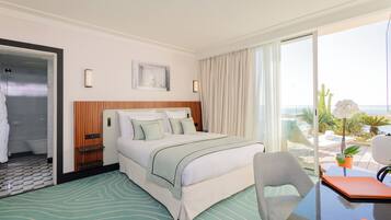 A hotel room with a bed, a chair, a table, a bathroom, and a balcony with plants.
