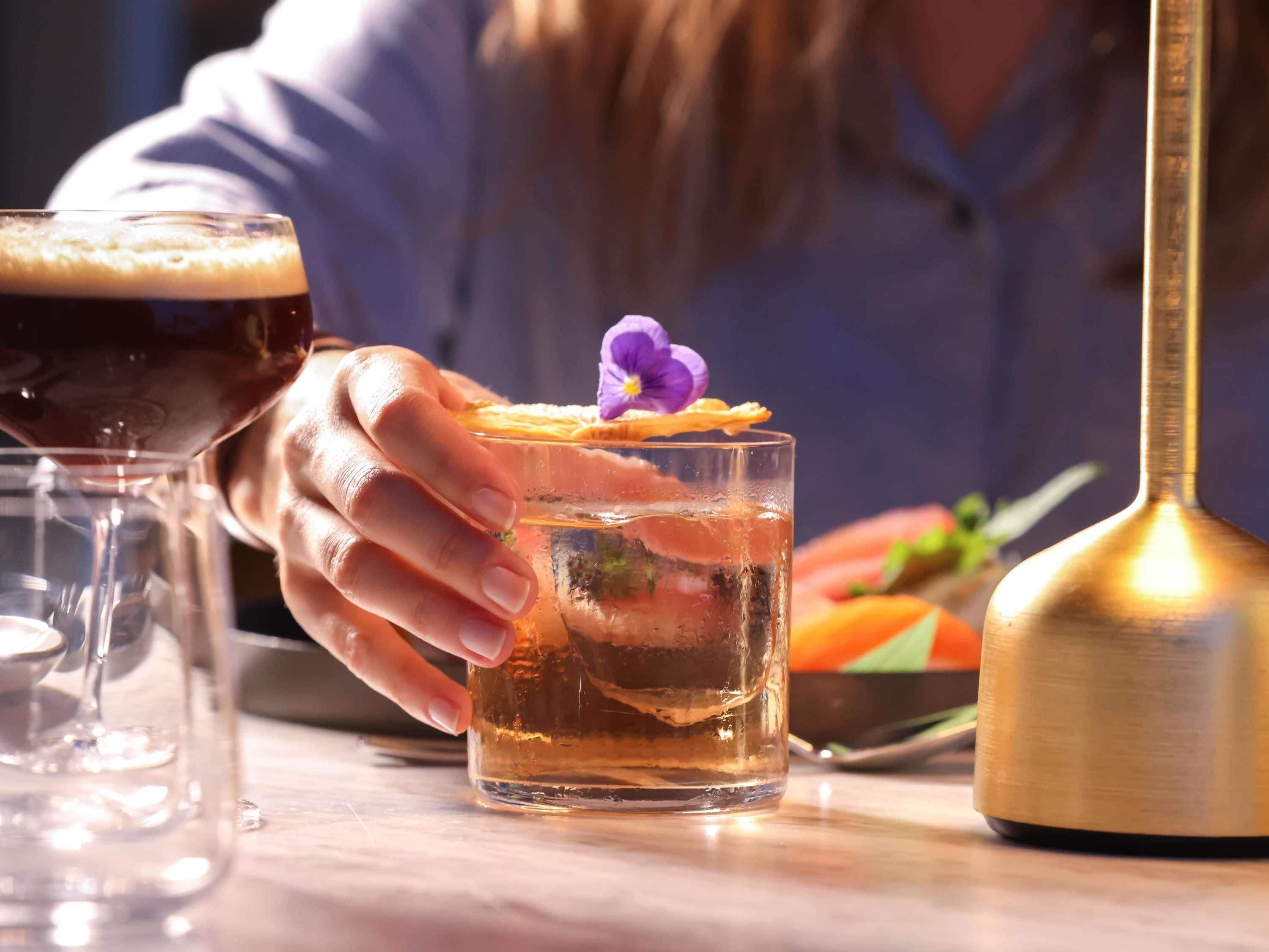 A person holding a cocktail glass with a garnish, a bottle, and a glass of red drink on a table.
