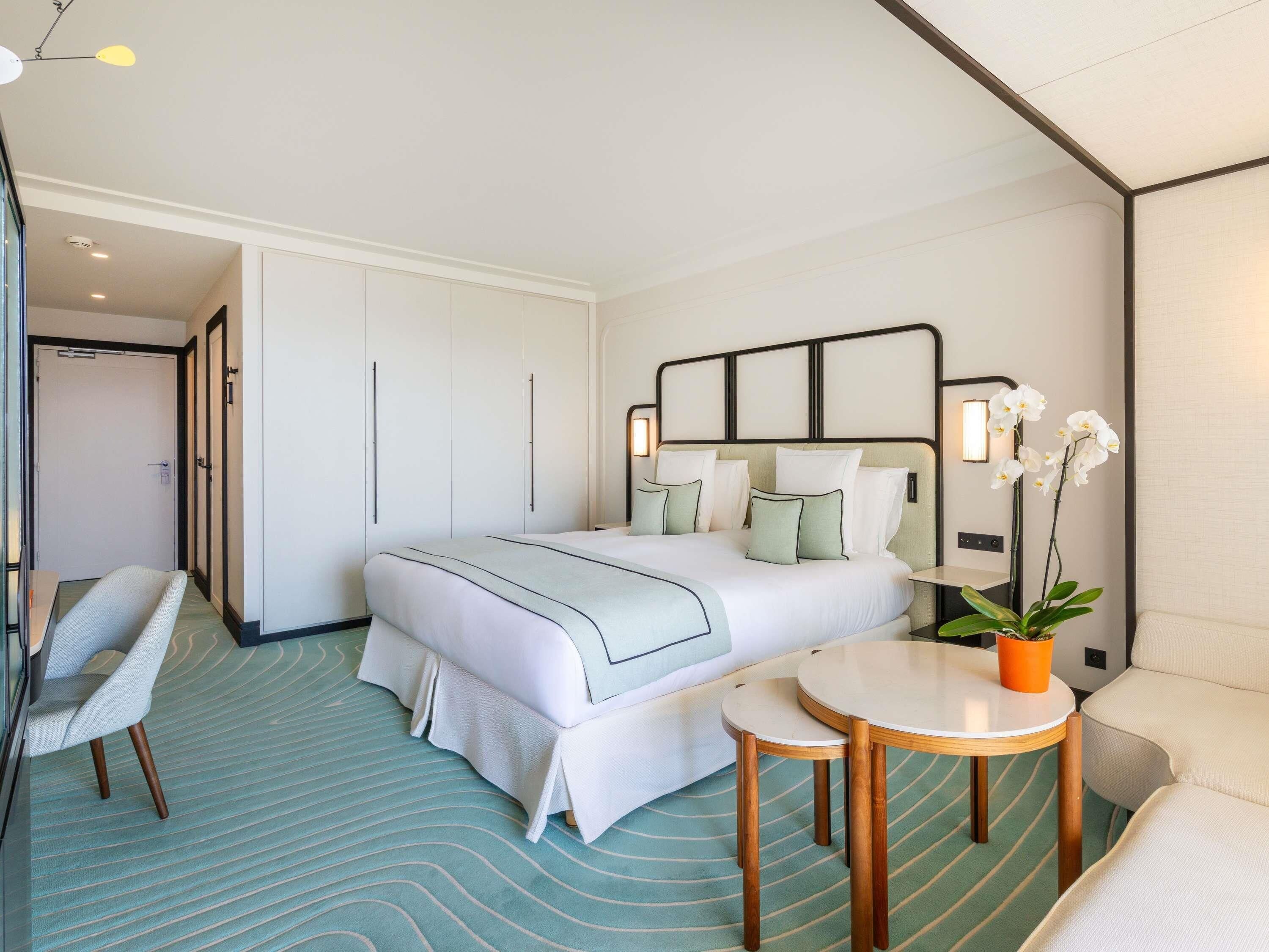 A hotel room with a bed, bedside table, chair, and a potted plant.