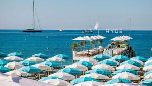 A beachside hotel with rows of sun umbrellas and lounge chairs, a pier with a flag, and boats in the background. - Mondrian Cannes (Cannes)