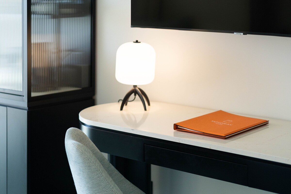 A modern hotel room with a desk, a chair, a lamp, and a book on the desk.
