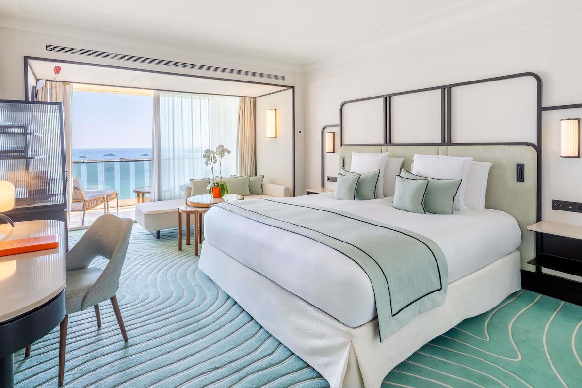 A hotel room with a bed, a desk, a chair, a small table with a plant, and a view of the ocean.