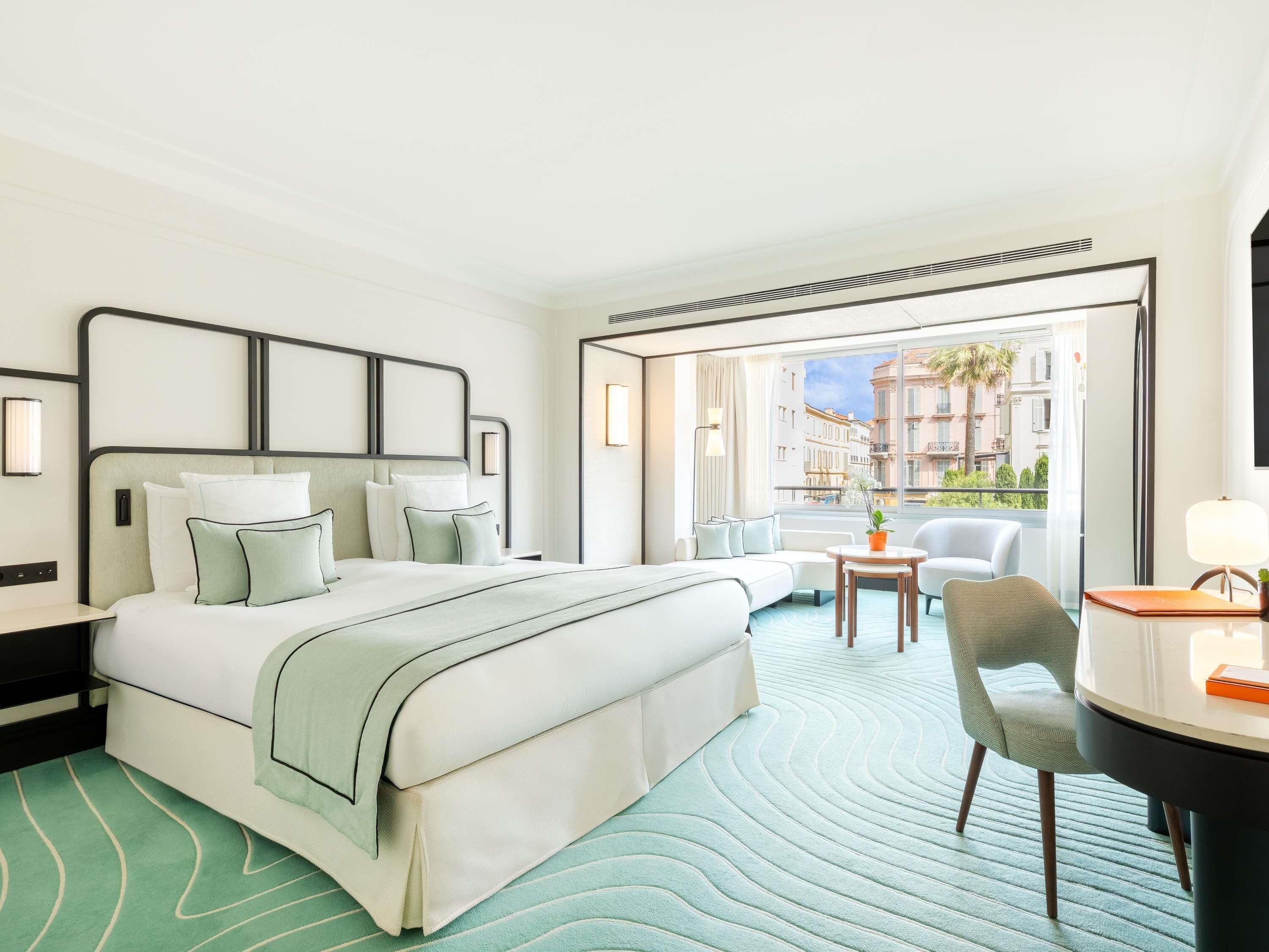 A hotel room with a bed, a desk, a chair, a small table, and a view of the city.