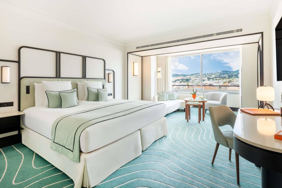 A hotel room with a large bed, a desk, a chair, and a view of a cityscape.