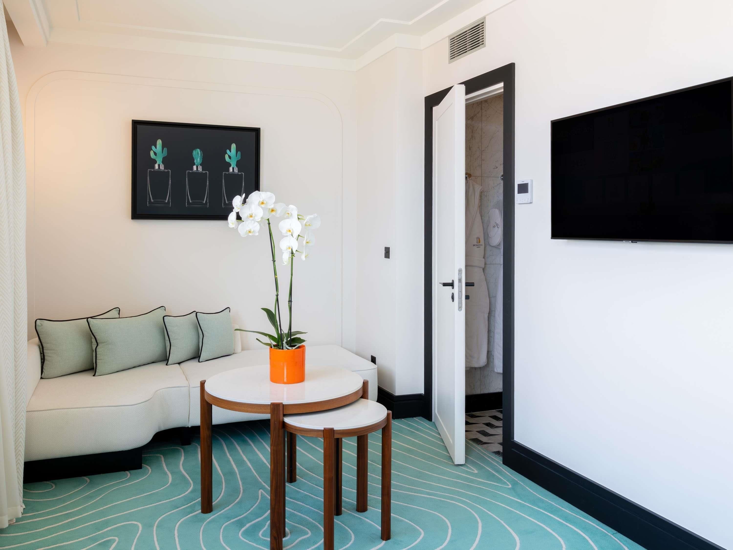 A hotel room with a sofa, a small table with a potted orchid, a framed picture on the wall, and a TV mounted on the wall.