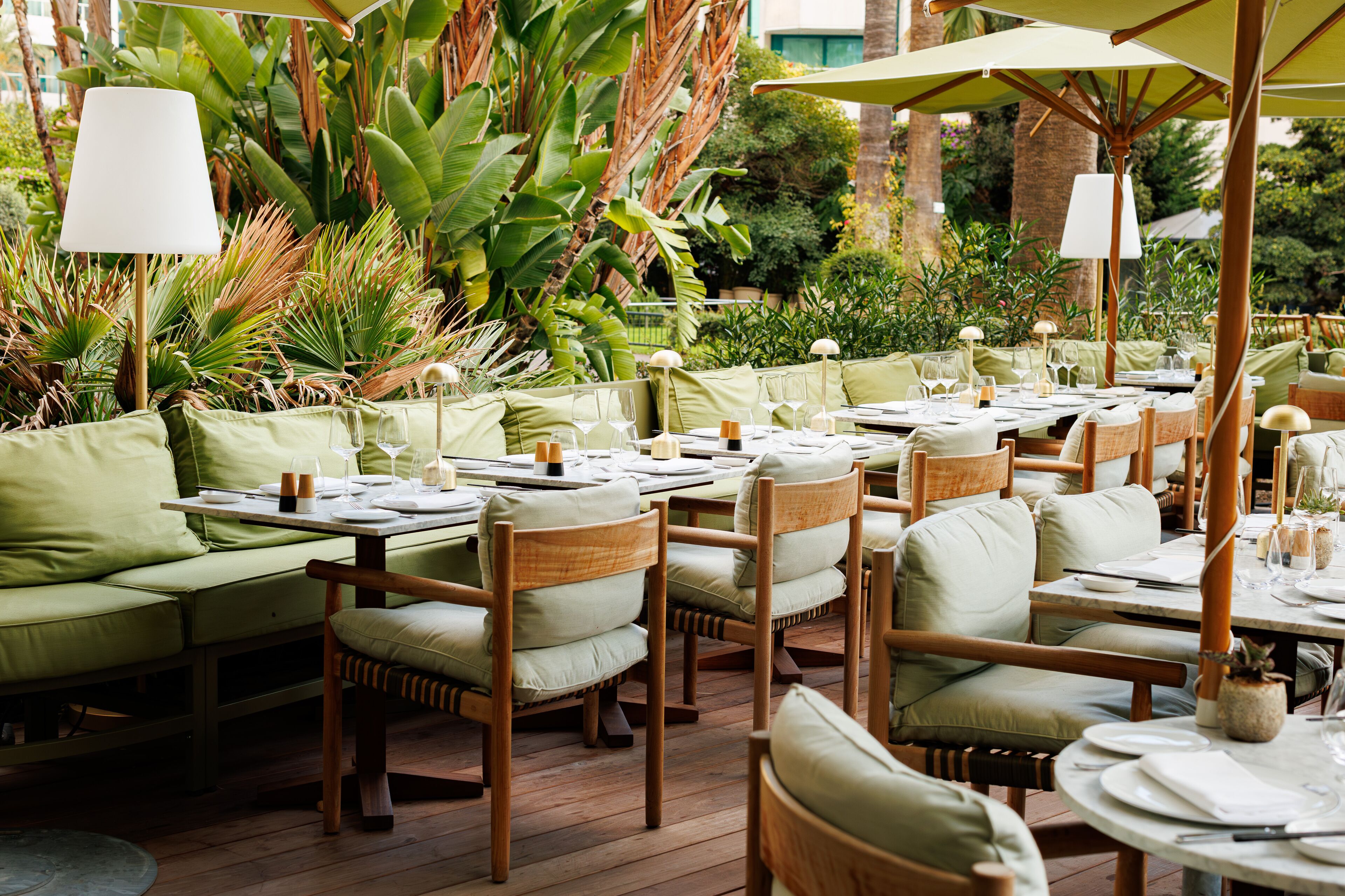 Un área de comedor al aire libre con mesas y sillas de madera, rodeada de abundante vegetación y plantas.