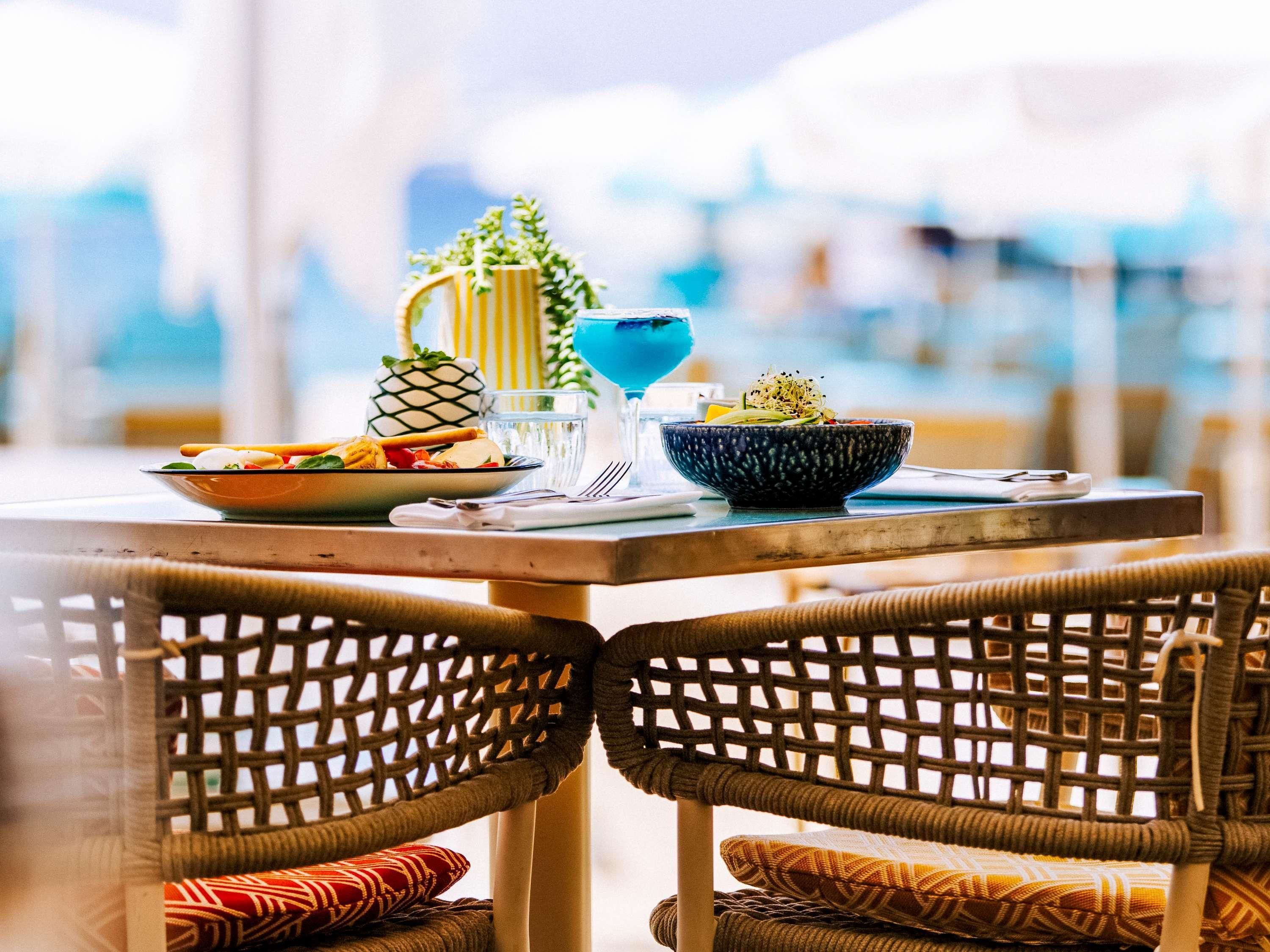 Une table à manger avec un saladier rempli de salade, un ananas et une boisson bleue. Des chaises en osier avec des coussins sont disposées autour de la table.