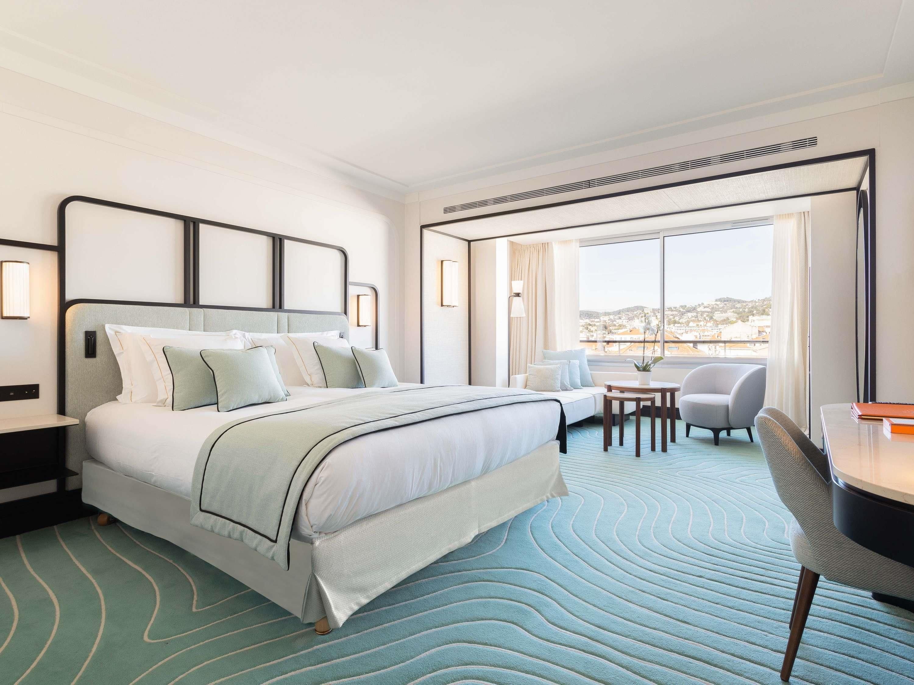 A modern hotel room with a large bed, a small table with chairs, and a view of the city through large windows.