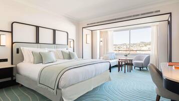 A modern hotel room with a large bed, a small table with chairs, and a view of the city through large windows.