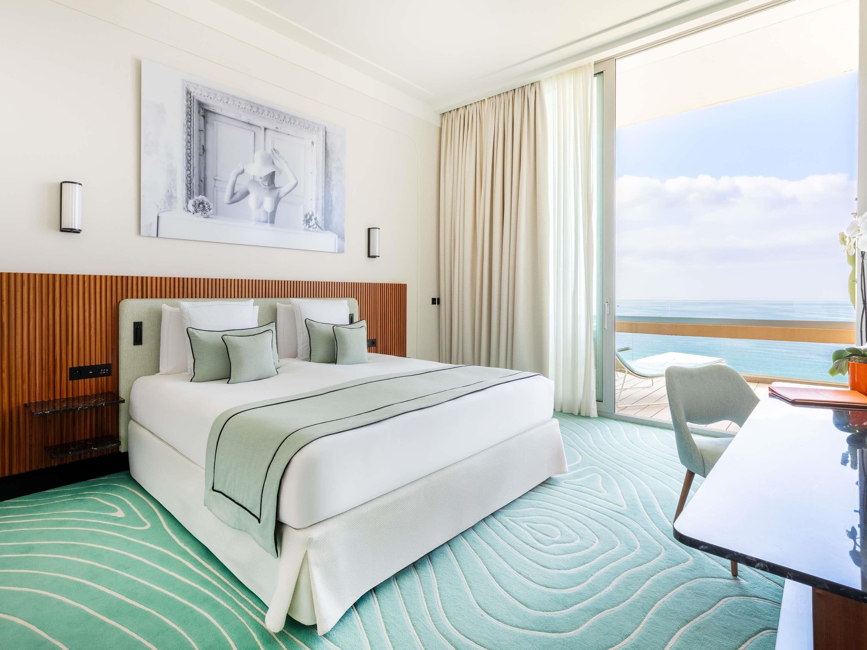 A hotel room with a bed, a desk, a chair, a view of the ocean, and a painting on the wall.