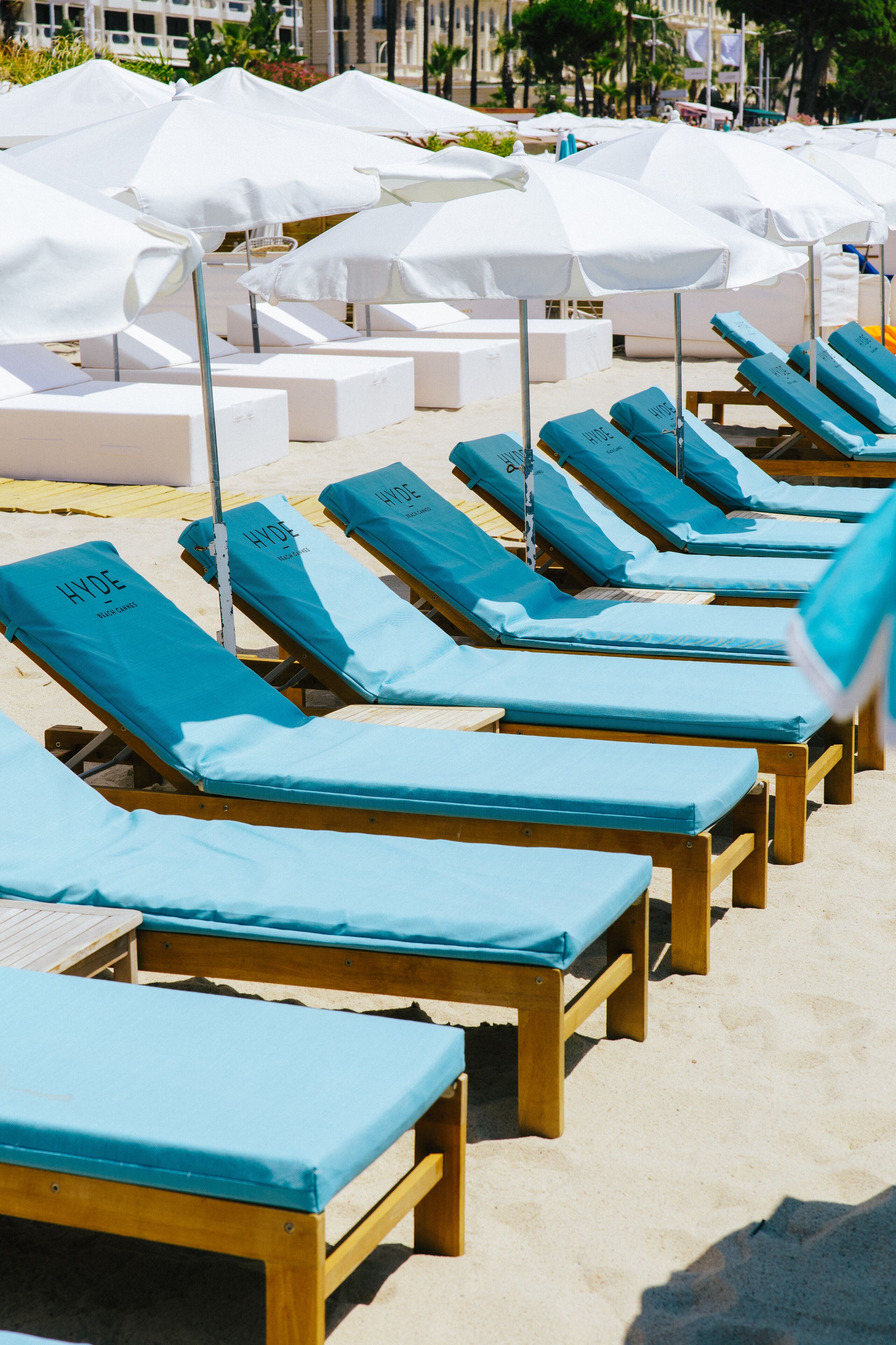 Rows of wooden lounge chairs with blue cushions and white umbrellas on a sandy beach.