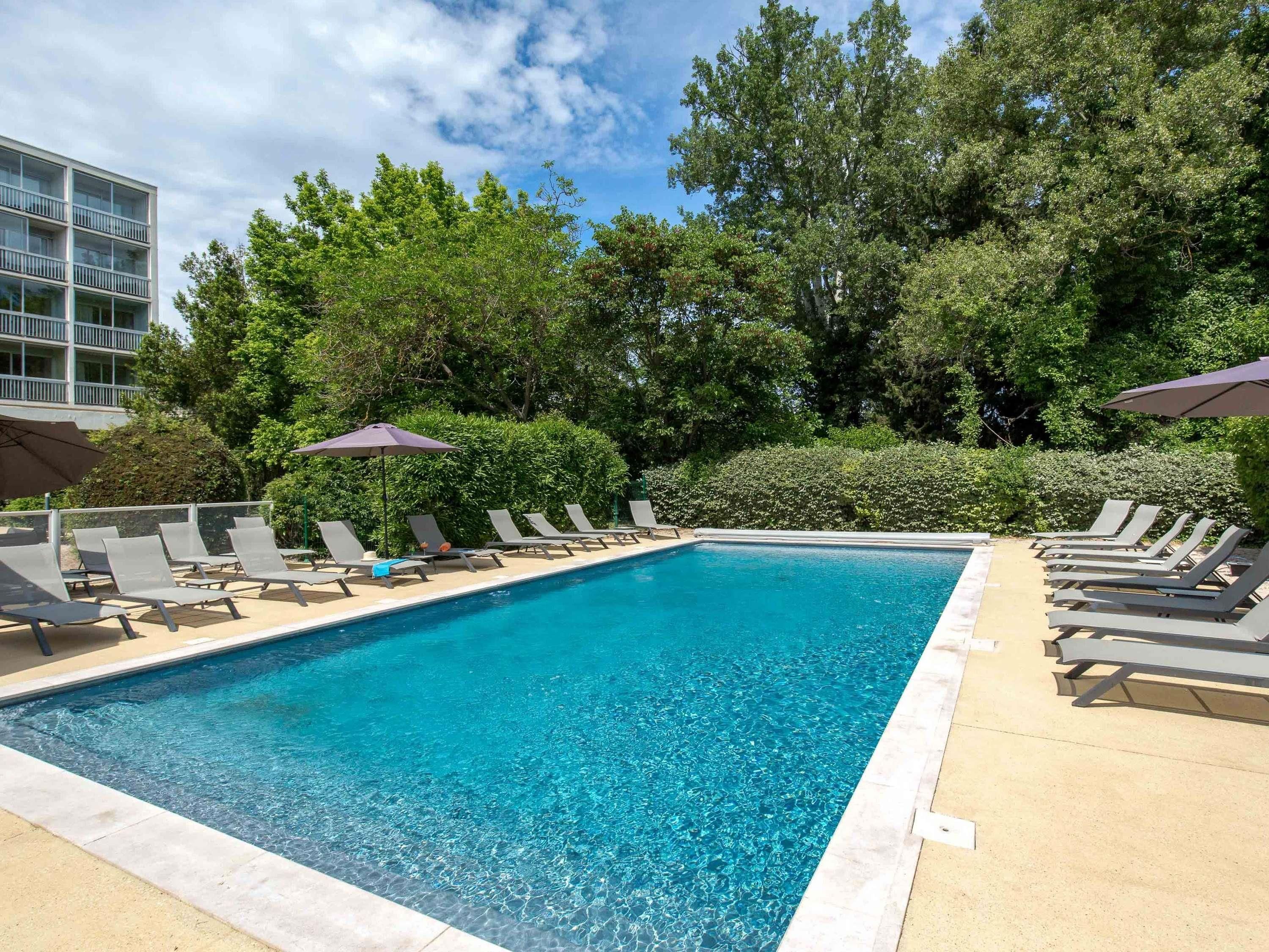 Una piscina rectangular con agua azul cristalina, rodeada de sillones de descanso y sombrillas, con árboles y un edificio al fondo.
