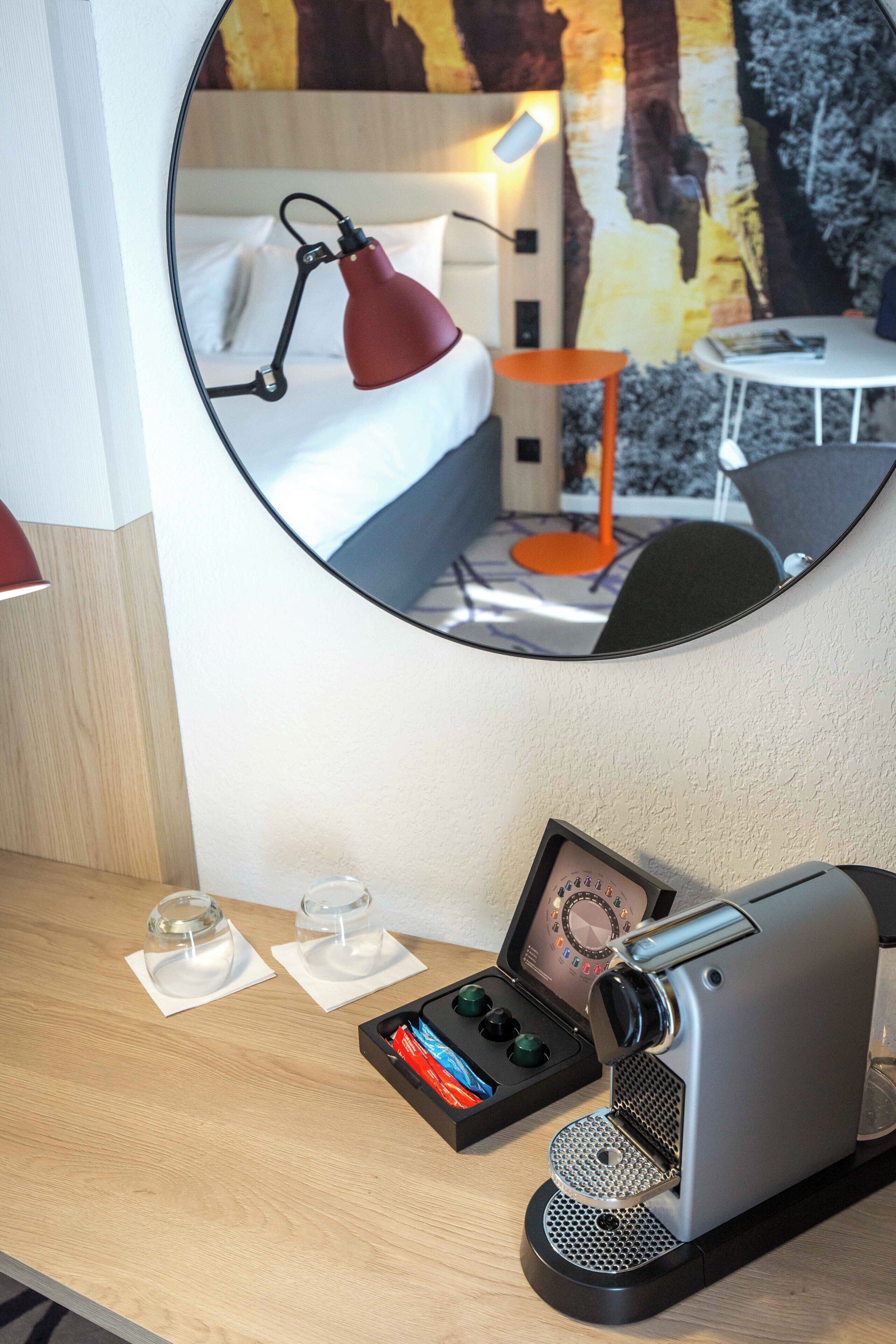 Ein Hotelzimmer mit einem runden Spiegel, der ein Bett, eine rote Schreibtischlampe, eine Kaffeemaschine und einen kleinen Tisch mit zwei Gläsern reflektiert.