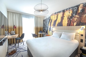 Habitación de hotel con una cama grande, un escritorio con silla, una mesita redonda pequeña y un cuadro grande en la pared.