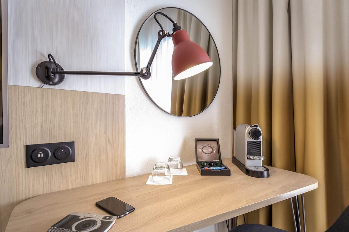 A modern hotel room with a wooden desk, a red desk lamp, a coffee machine, and a mirror.