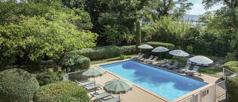 Piscina rectangular con sillas de descanso y sombrillas, rodeada de setos y árboles cuidados.